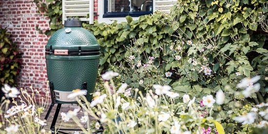 Donkergroene Big Green Egg barbecue op een standaard in een tuin met bloemen en planten.
