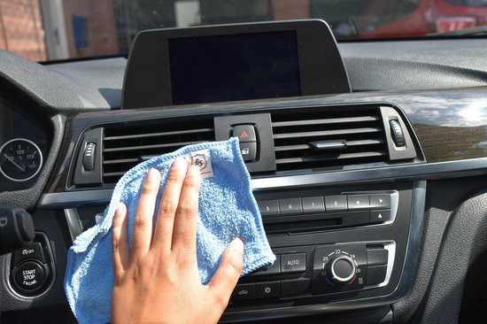 Hand gebruikt een blauw microvezel brillendoekje om het dashboard van een auto schoon te maken.