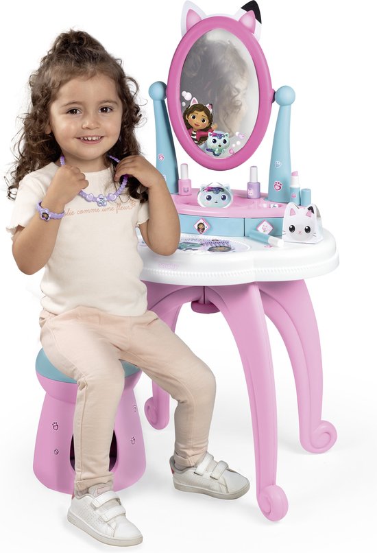 Meisje met krullend haar zit op krukje met Gabby's Dollhouse kaptafel, met ketting en armband.