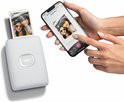 Handen houden een smartphone vast met een Instax foto-app. Naast de smartphone een witte Instax Mini Link 2 printer, met een foto van twee lachende personen die naar de camera kijken.