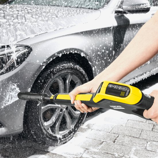Een hand met een gele Kärcher hogedrukreiniger spuit water op een grijze auto met veel schuim.