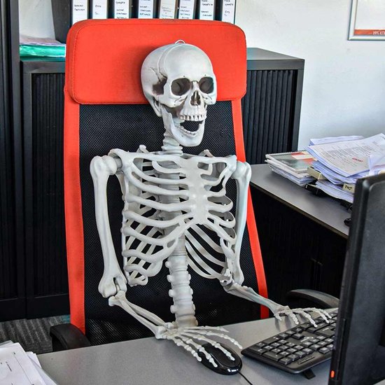 Een levensgroot skelet zit achter een bureau aan een computer in een kantoorstoel.