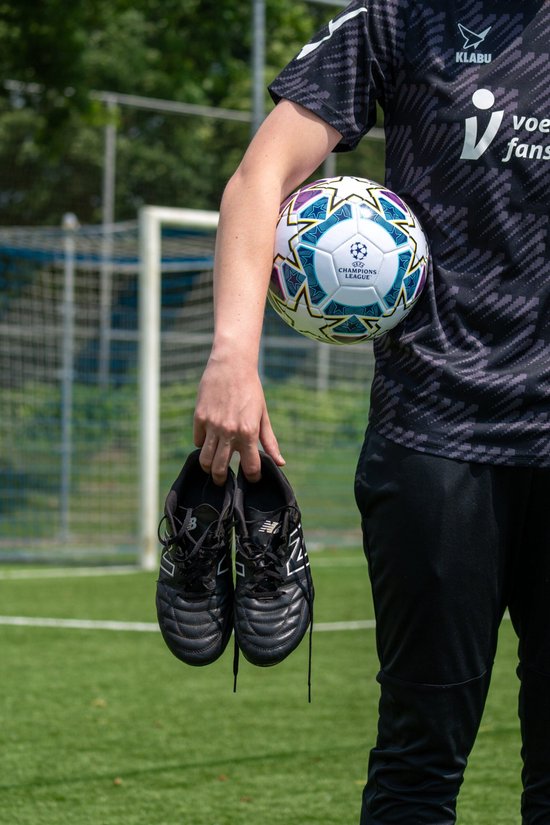 Persoon in sportkleding houdt een voetbal met UEFA Champions League logo vast. De persoon draagt een voetbalshirt met tekst en houdt schoenen vast.