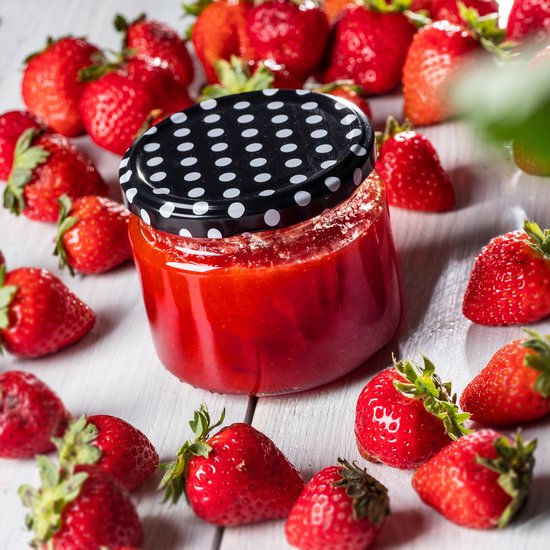 Bocal de confiture de fraises avec un couvercle à pois noirs, entouré de fraises fraîches.
