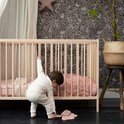 Baby bukt bij een ledikant. Draagt een beige broekpak.