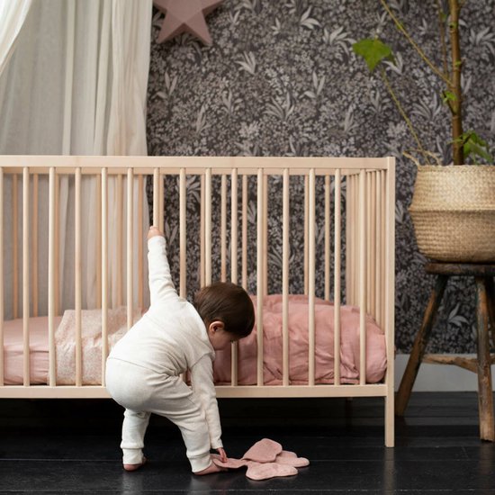 Baby bukt bij een ledikant. Draagt een beige broekpak.