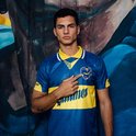 Man model wijst naar het wapen op het blauwe voetbalshirt met gele accenten en de tekst 'Quilmes'.