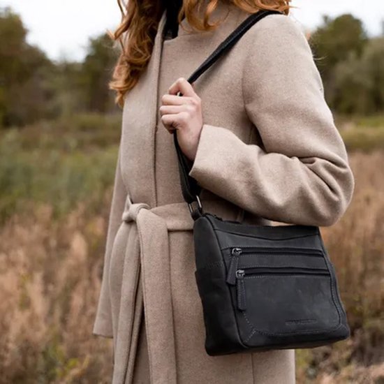 Zwarte leren schoudertas met ritsvakken, gedragen door een vrouw in een beige jas.