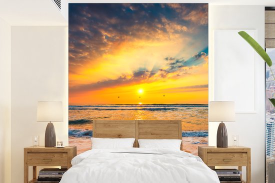 Fotobehang van een zonsondergang op zee met oranje, gele en blauwe wolken, golven op het strand.