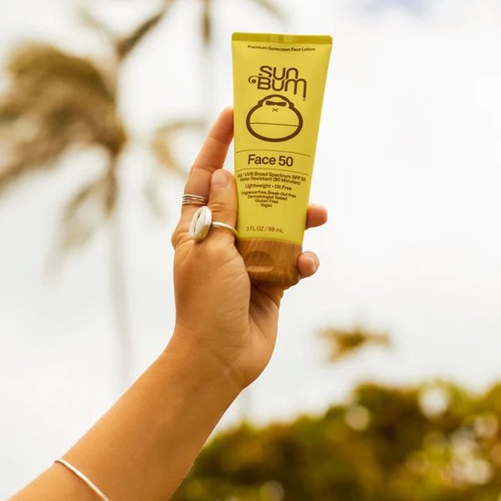 Main tenant un tube jaune de crème solaire Sun Bum Face 50, palmier en arrière-plan.