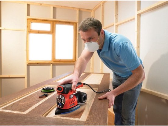 Man met mondkapje schuurt met een oranje en zwarte BLACK+DECKER multischuurmachine op een houten deur.