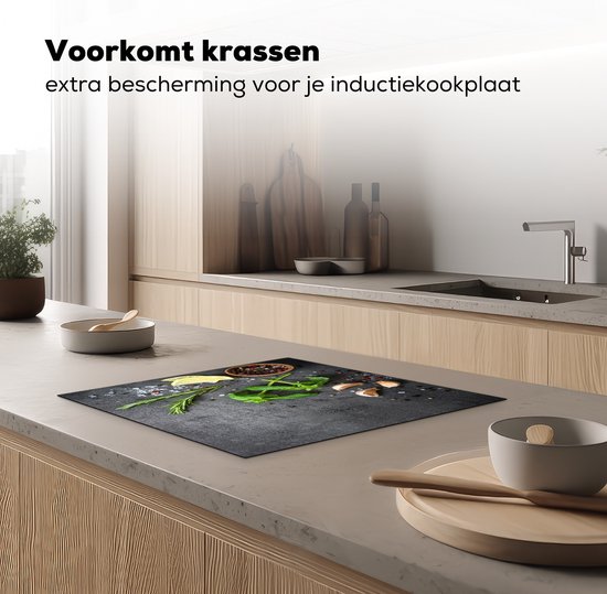 Zwarte inductiebeschermer met betonprint, bedekt met kruiden, knoflook, citroen en peperkorrels, op een aanrecht in een moderne keuken.