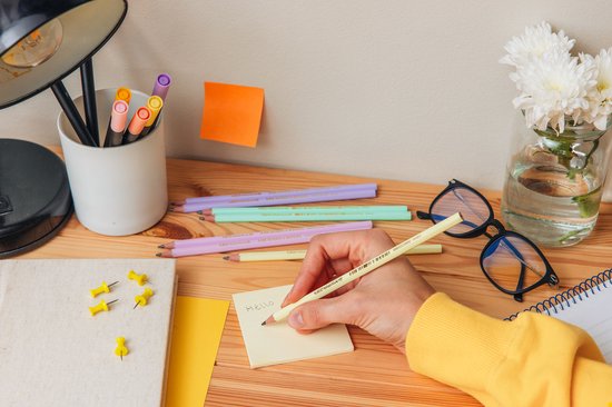 Hand schrijft 'Hello' met een lichtgeel potlood, bril, bloemen en potloden op bureau.