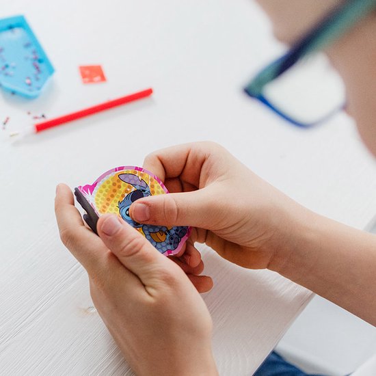 Un enfant tient un magnet Stitch diamond painting presque terminé. Le magnet montre Stitch sur fond jaune, partiellement recouvert de strass.