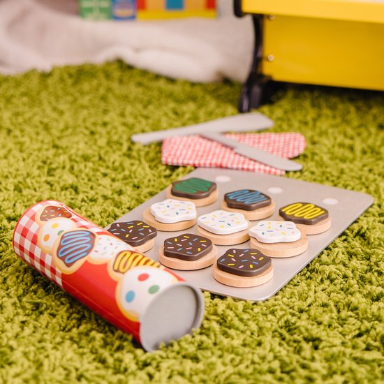 Houten koekjes op bakplaat, met rol koekjesdeeg, ovenwant en spatels op groen tapijt. Koekjes met witte icing, sprinkles, chocolade, groene en blauwe icing.