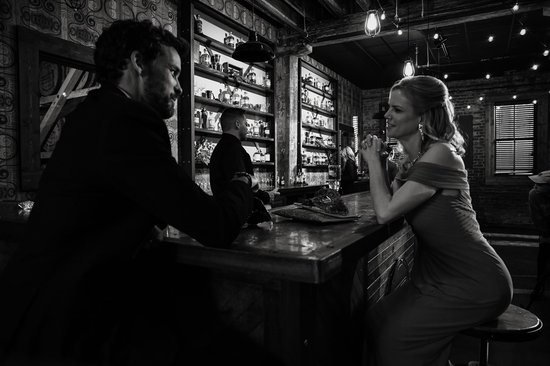 Man in pak en vrouw in rode jurk aan een bar, pratend met een barman op de achtergrond.