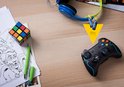 Zwarte game controller, koptelefoon, Rubik's kubus en stripboek op een houten bureau met twee AAA Varta batterijen.