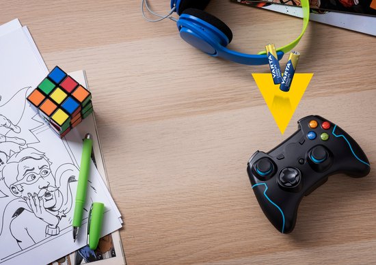 Zwarte game controller, koptelefoon, Rubik's kubus en stripboek op een houten bureau met twee AAA Varta batterijen.