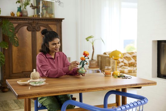 Une femme assise à une table construit un chrysanthème Lego. Les fleurs sont orange et rouges.