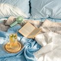 Een open boek, een glas ijsthee en een ventilator liggen op blauwe, grijze en witte wafelstof dekens.