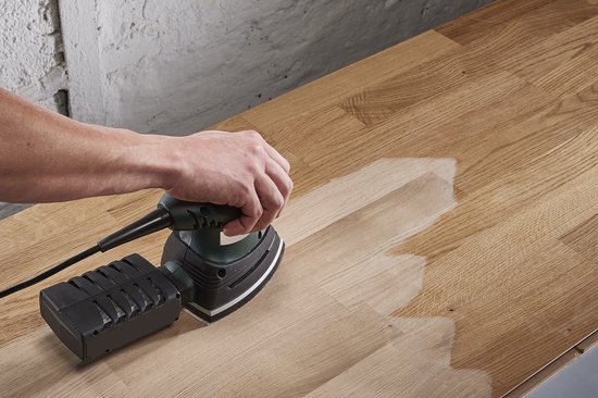 Een hand houdt een groen-zwarte multischuurmachine vast op een houten tafel.