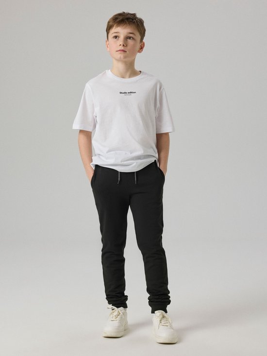 Jongen in wit T-shirt en zwarte joggingbroek, staand met handen in de zakken.