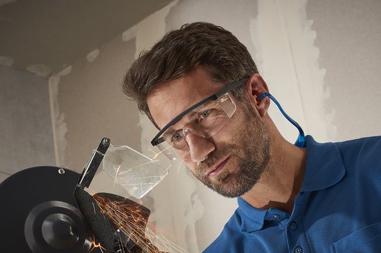Man met blauwe polo draagt gehoorbescherming met oranje oordopjes en veiligheidsbril, terwijl hij bij een slijptol staat.