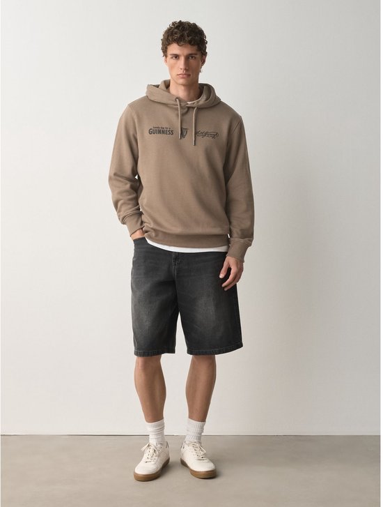 Man in bruine hoodie met opdruk 'GUINNESS' op de borst, zwarte spijkerbroek en witte sneakers.