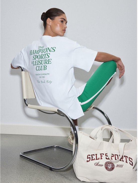 Model zit op een stoel en draagt een wit T-shirt met groene tekst 'THE HAMPTONS SPORTS & LEISURE CLUB' op de rug. Naast haar staat een beige tas met tekst.