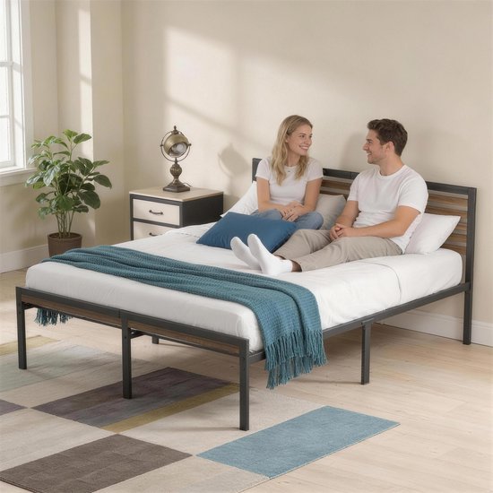 Koppel zittend op een bed met zwart metalen frame en houten hoofdbord, in een kamer met een blauw kussen en sprei.