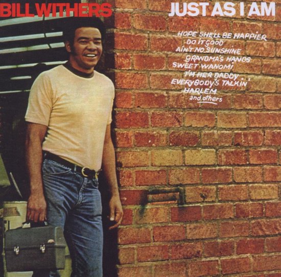 Bill Withers staat voor een bakstenen muur, hij draagt een geel shirt en jeans en houdt een aktetas vast.