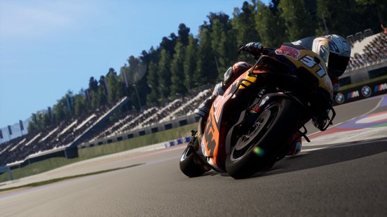 Coureur in oranje en zwart pak op een motor, bocht op circuit met tribunes en bomen op de achtergrond.