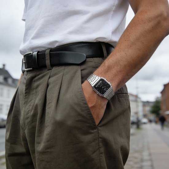 Bras portant une montre argentée avec bracelet métallique, cadran noir, heure analogique et numérique, avec un t-shirt blanc et un pantalon marron.