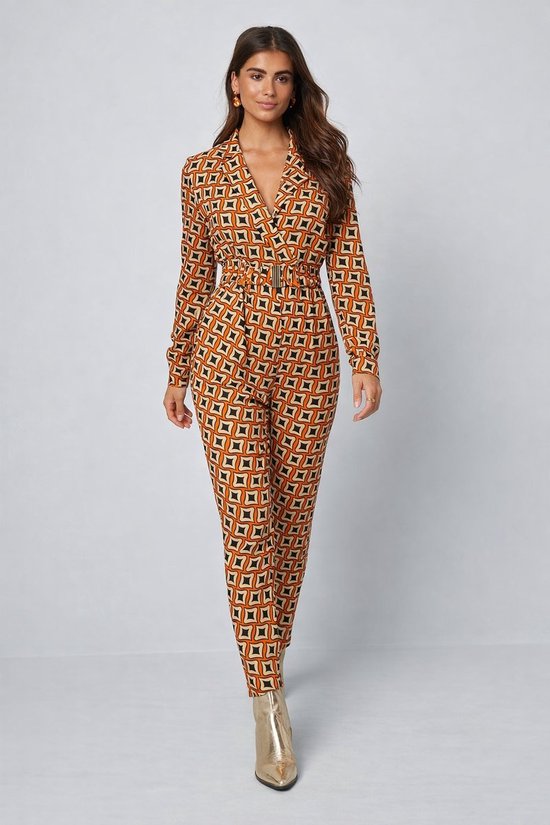 Jumpsuit met geometrisch oranje, zwart en beige patroon, lange mouwen, riem met gesp en goudkleurige laarzen. Model draagt de jumpsuit.