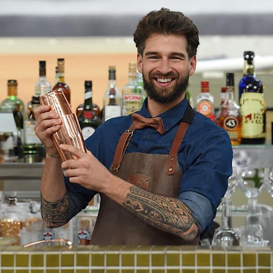 Man met tatoeages in een blauw overhemd, schort en roestoranje strik, schudt een koperen cocktailshaker.