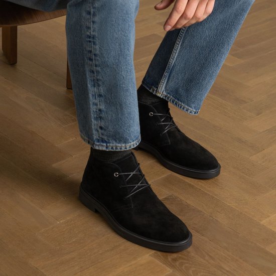 Homme assis sur une chaise, portant un jean et des bottes à lacets en daim noir.