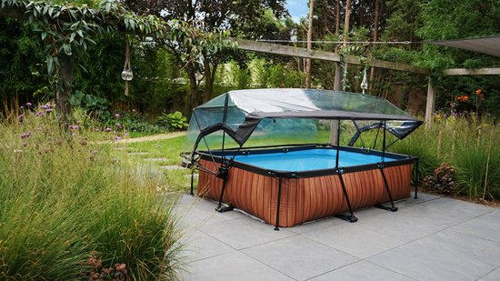 Rechthoekig zwembad met houtlook bekleding en zwarte rand, met transparante opzetzwembad overkapping, in een groene tuin op een betegeld terras.