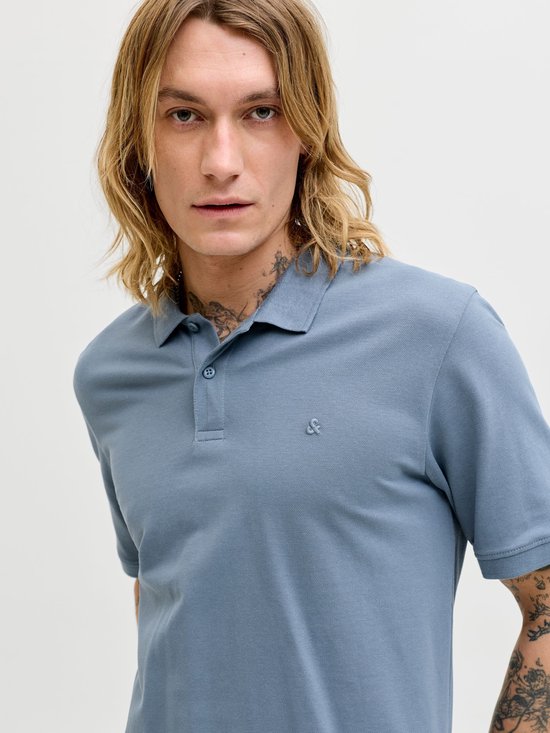 Close-up van een man met lang blond haar en tatoeages, draagt een blauw poloshirt.