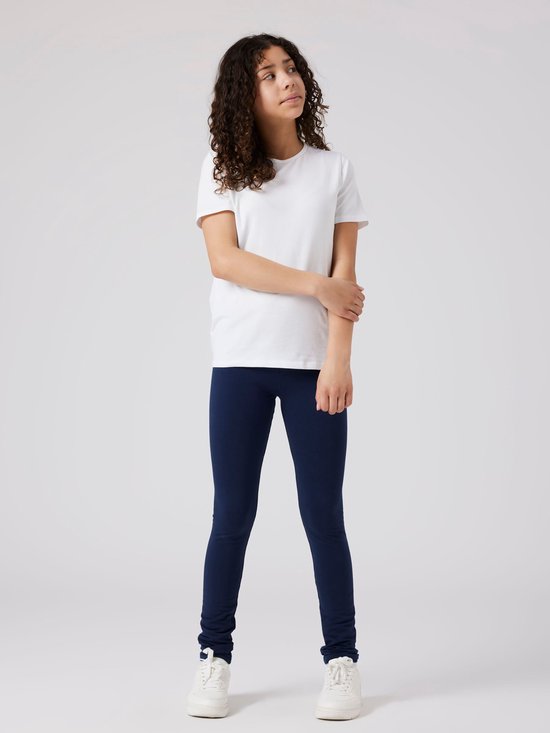 Meisje met krullend haar, een witte T-shirt en donkerblauwe legging, staand.