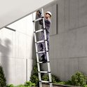 Man in overall en helm werkt met boormachine aan een gevel, staand op een opengevouwen ladder.