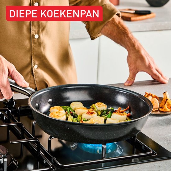 Man kookt sint-jakobsschelpen en groenten in een diepe zwarte koekenpan op een gasfornuis.