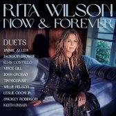 Rita Wilson zittend op een bed in een glinsterende zwarte jurk, met de tekst "Rita Wilson Now & Forever Duets" en een lijst met artiesten.