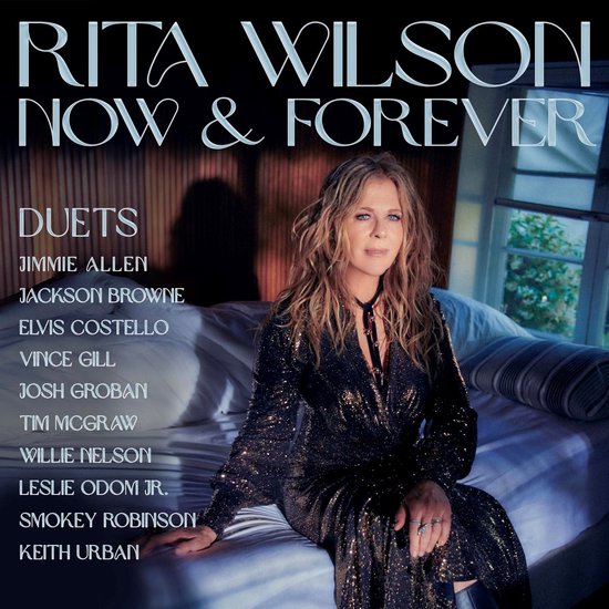 Rita Wilson zittend op een bed in een glinsterende zwarte jurk, met de tekst "Rita Wilson Now & Forever Duets" en een lijst met artiesten.