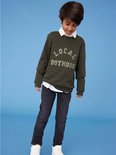 Jongen met witte overhemdkraag en legergroene sweater met tekst, draagt een grijze slim fit spijkerbroek.