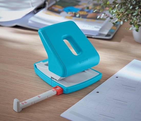 Lichtblauwe Leitz perforator met witte en rode details, staand op een houten bureau naast vellen papier.