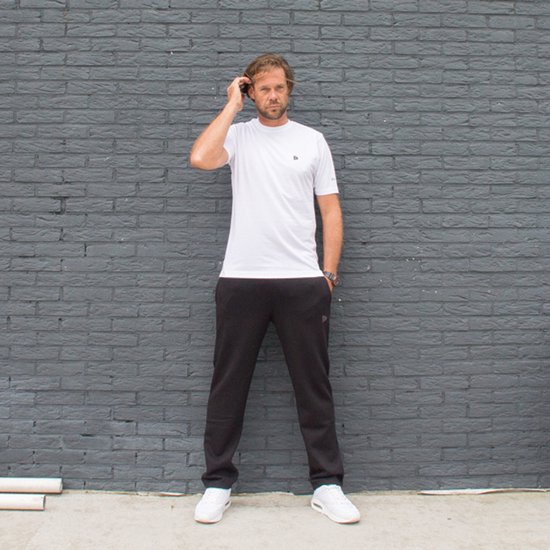 Man in wit Donnay t-shirt, zwarte broek en witte sneakers staat voor donkergrijze bakstenen muur.
