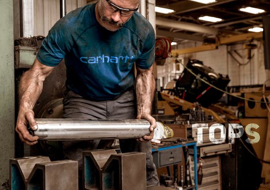 Blauwe Carhartt werkshirt met tekst op de borst, gedragen door een man met een snor en veiligheidsbril in een werkplaats.