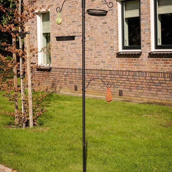 Zwart vogelvoederstation naast een bakstenen muur met twee voerbakjes, een netje met vogelvoer en een peer-vormig vogelvoer.