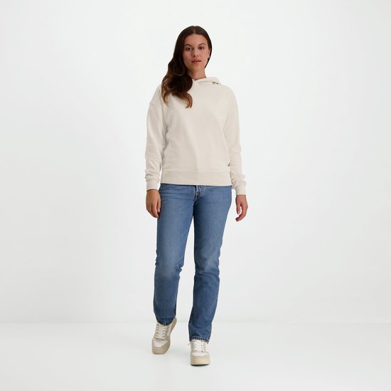 Vrouw met bruin haar, gekleed in een crème hoodie en spijkerbroek, staat frontaal.