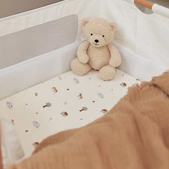 Drap de lit crème avec un imprimé de maisons, d'arbres et d'éléphants, avec un ours en peluche dessus.
